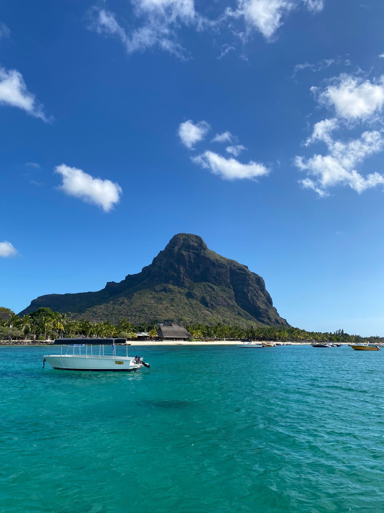Bright blue ocean with mountain backdrop, small boat, and clear sky with fluffy white clouds.