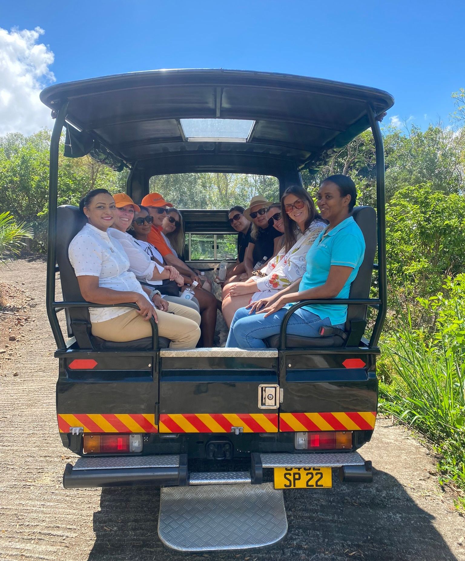 People riding in an open safari vehicle on a dirt road. Passengers are smiling. Sunny day.