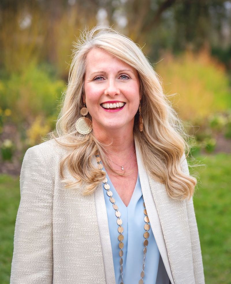 Woman with blonde hair, smiling outdoors, wearing a cream blazer, blue blouse, and gold jewelry.