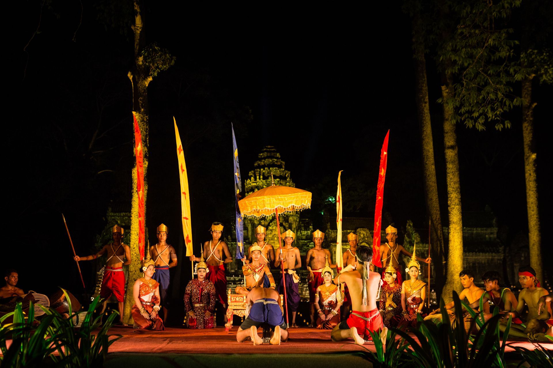 Group of performers on a brightly lit outdoor stage with colorful costumes and tall decorative banners