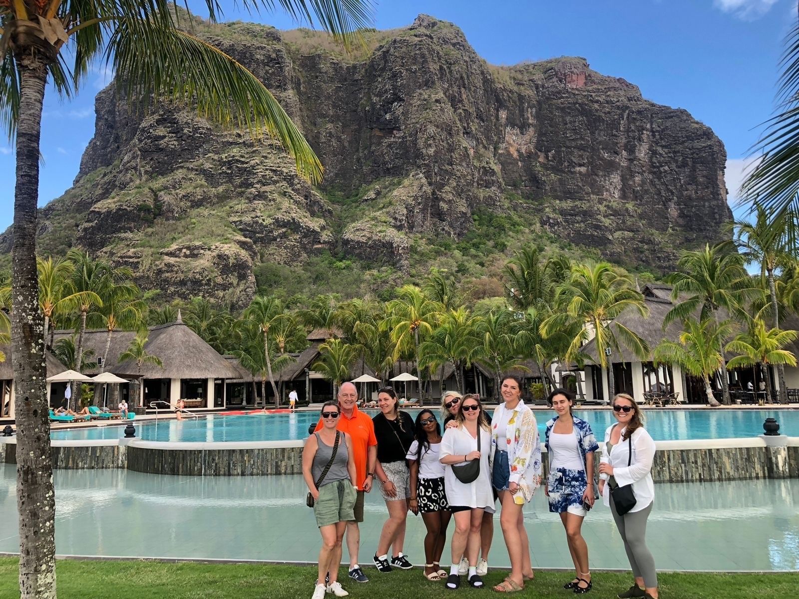 Group of people posing in front of a mountain, resort, and pool in Mauritius, sunny day.