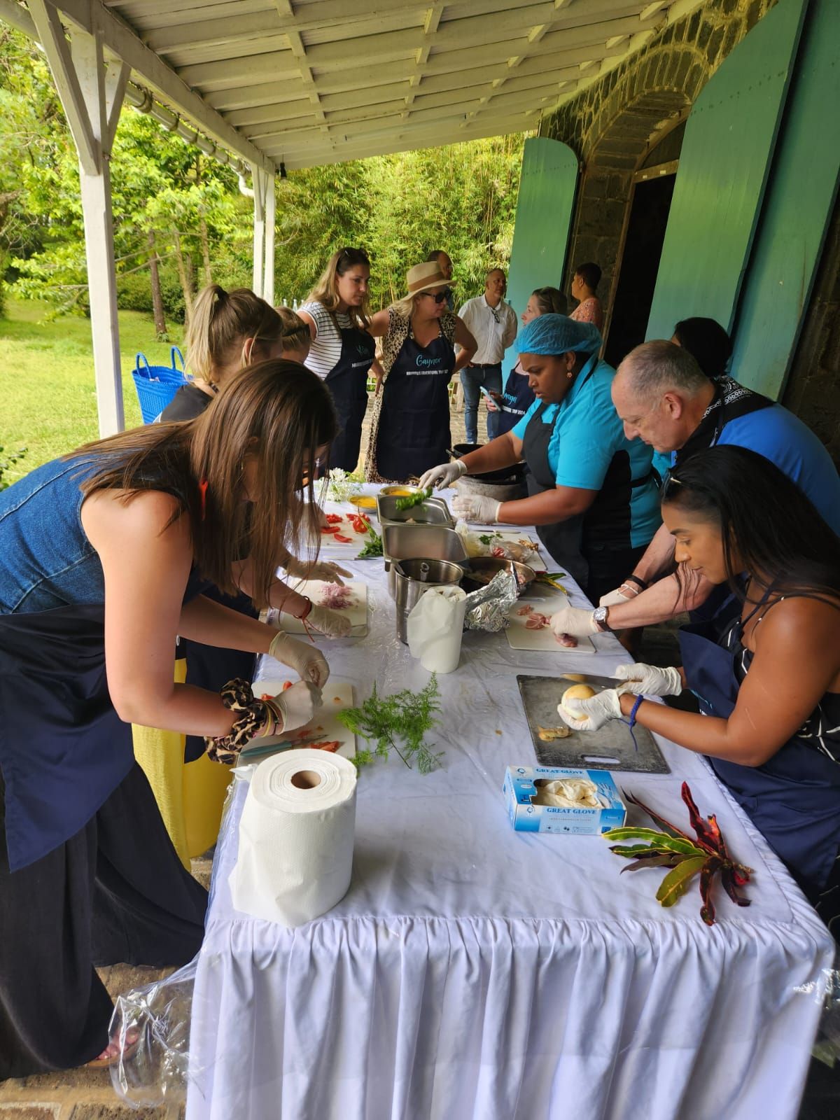 People preparing food at a table outside. They wear aprons and gloves, and ingredients are spread out.