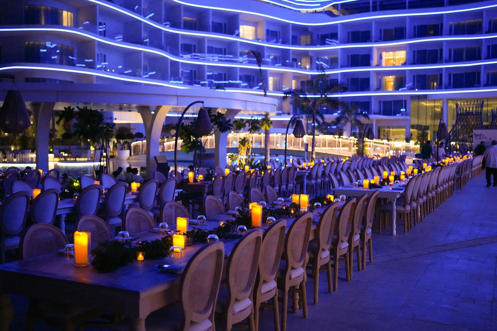 Outdoor evening event, tables set with candles, hotel in background, blue lighting.