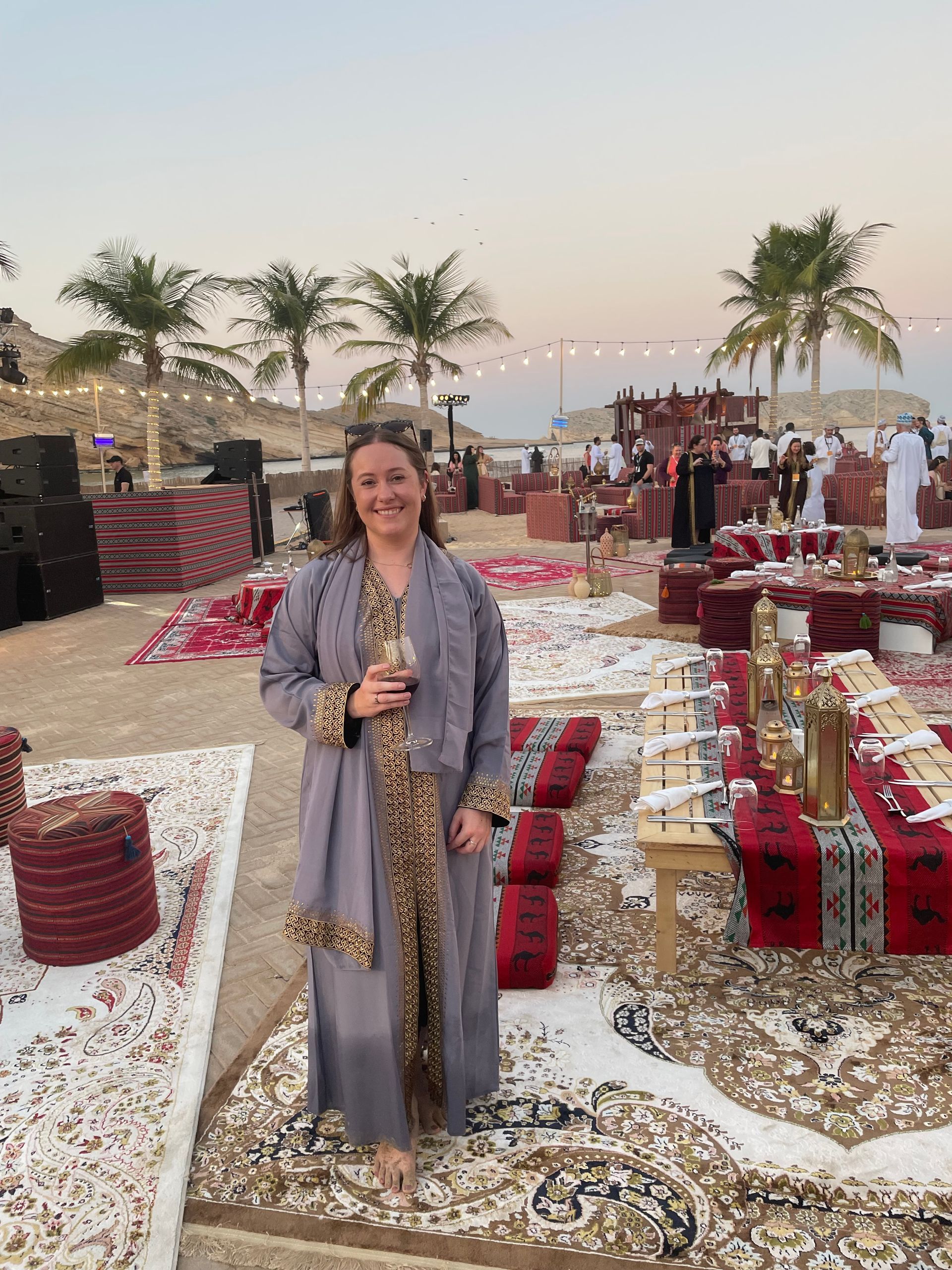 Woman in a gray robe with gold trim holding a glass, posing at an outdoor event with red carpets and palm trees.