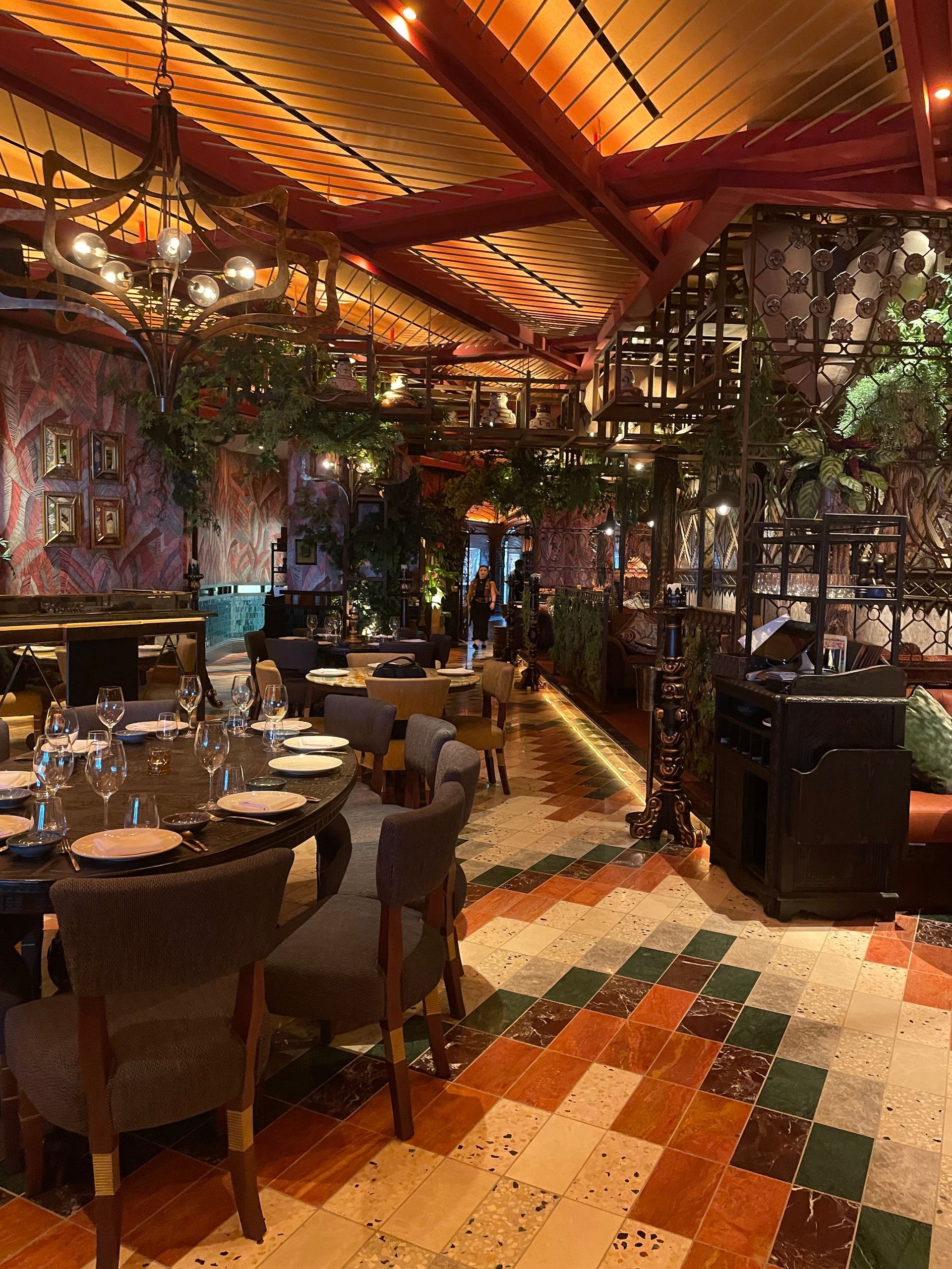 Restaurant interior: Long tables, patterned tile floor, lush greenery, ornate ceiling, and ambient lighting.