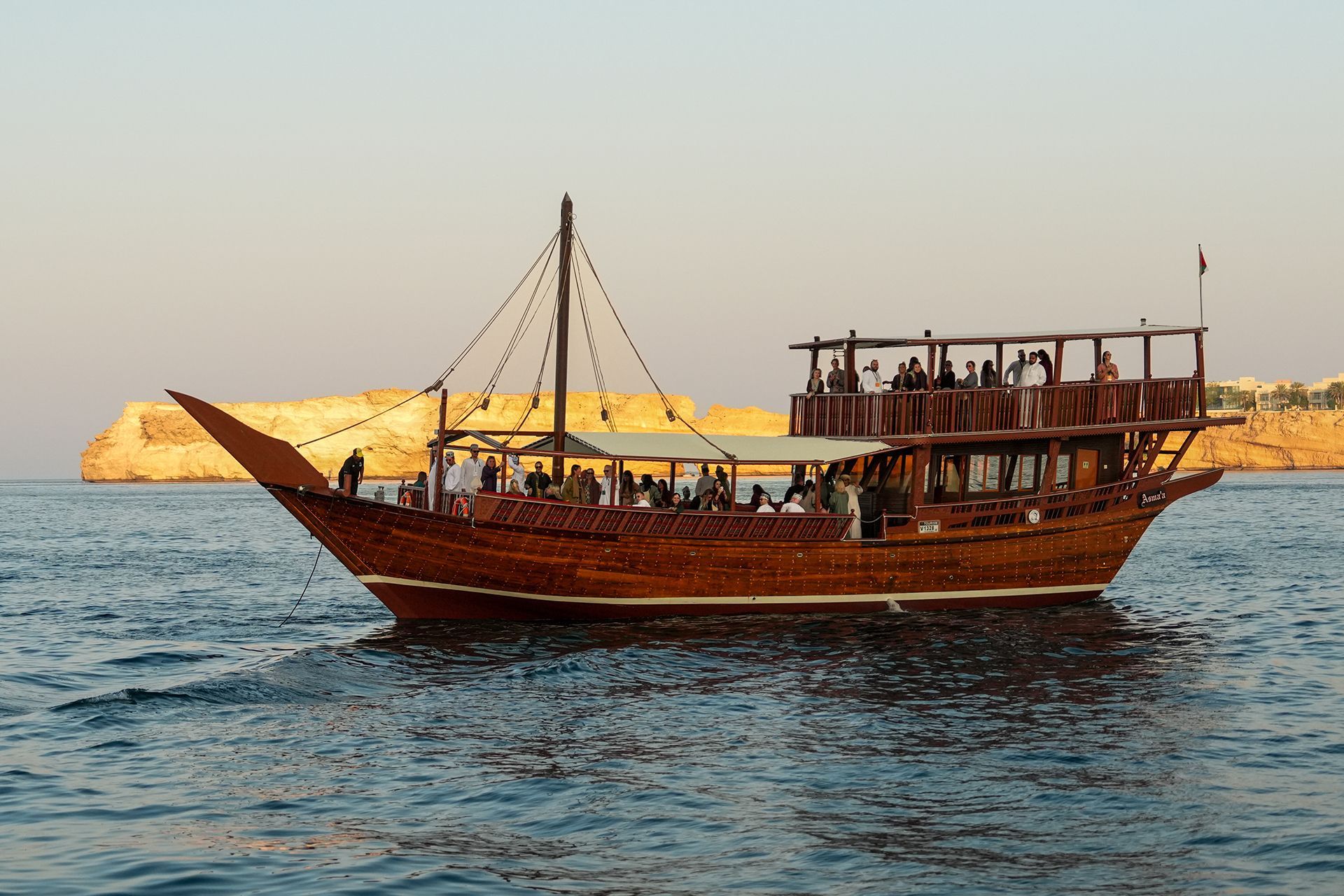 Wooden dhow sailing on water with people on board near rocky coastline at sunset.