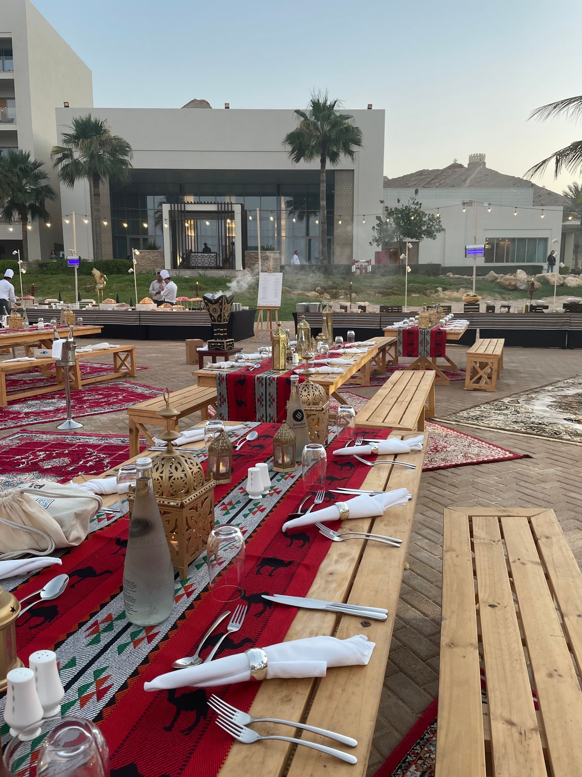 Outdoor dining tables set with red runners, lanterns, and silverware on a beach.