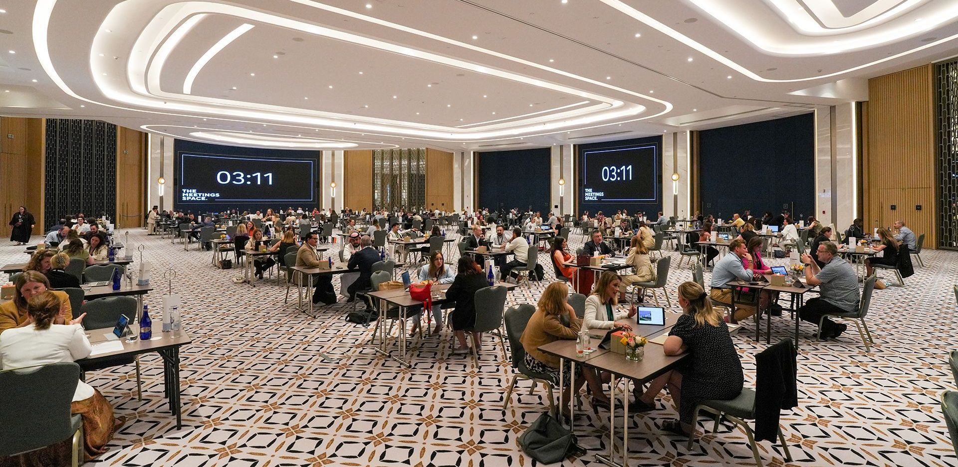 Large conference room with attendees seated at tables, large screens displaying text.