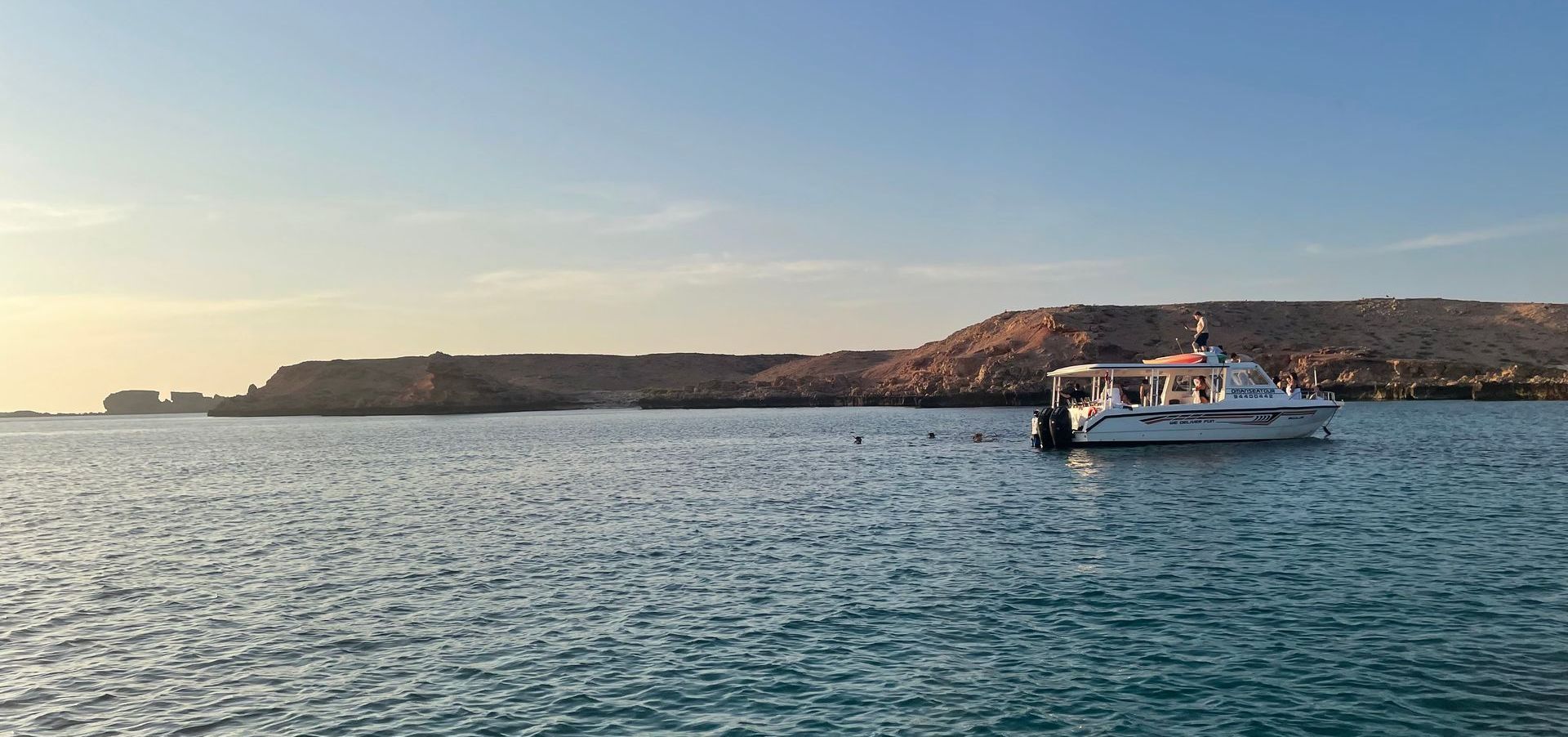 A white boat on the turquoise sea near a brown, rocky island under a clear blue sky.