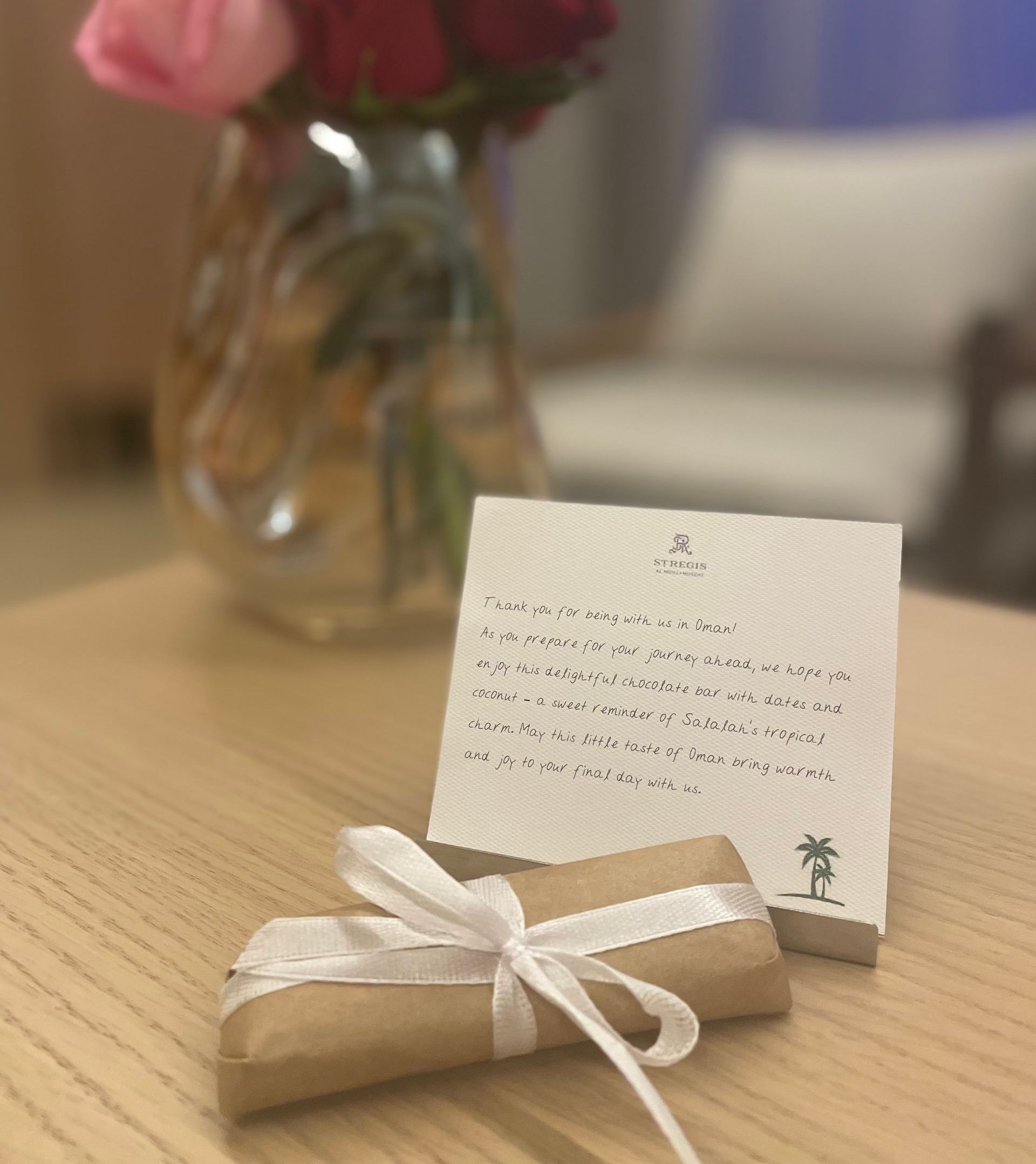 A gift wrapped in brown paper with a white ribbon and a handwritten note on a table; flowers and a blurred armchair in the background.