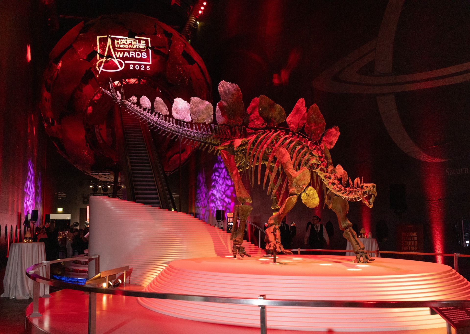 Stegosaurus skeleton on a red-lit stage at a museum event, with a large red sphere in the background.