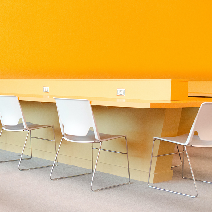 Three white chairs sit at a long, yellow desk against a bright yellow wall.