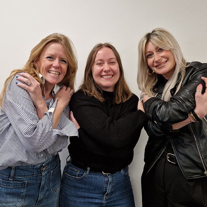 Three women smiling, hugging themselves in front of a white wall.