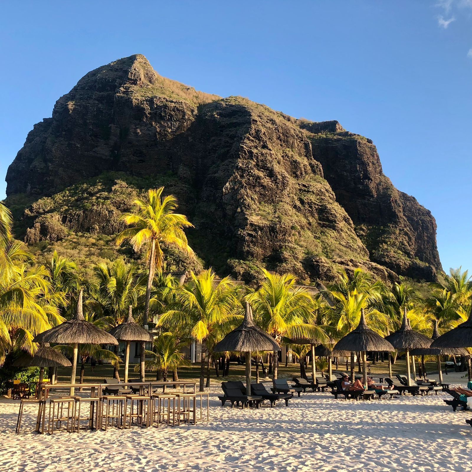 Beach scene with thatched umbrellas, palm trees, and a large, rocky mountain backdrop.