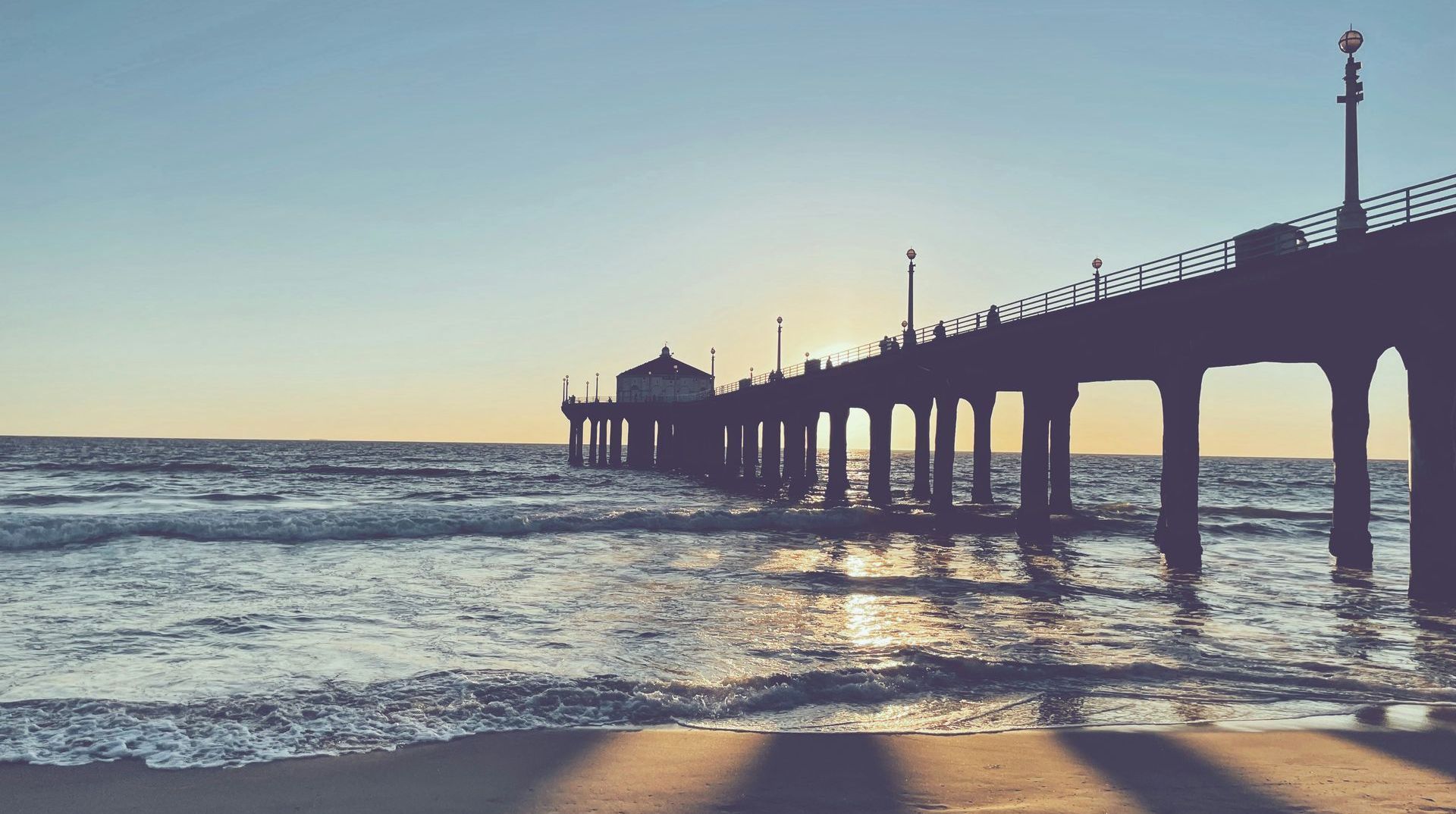 Sunset over a pier extending into the ocean; people silhouetted at the end, warm light reflecting on water.