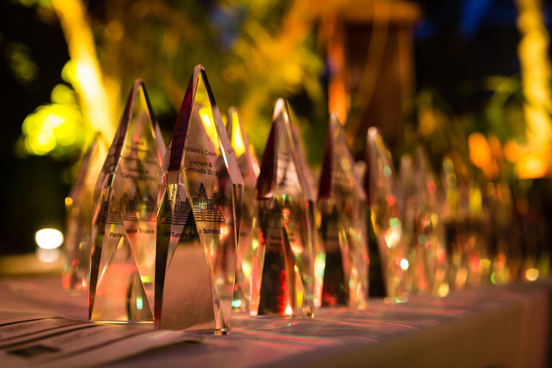 Awards lined up on a table, clear crystal shapes with etched text, outdoor setting with blurred lights.