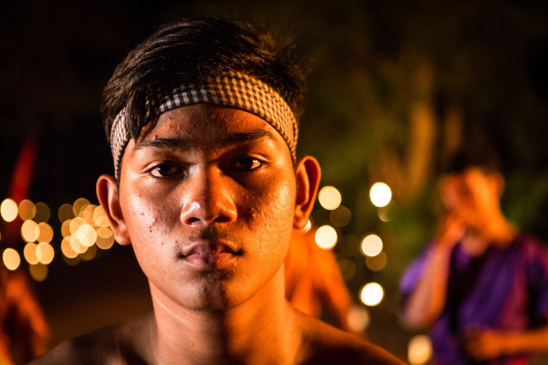 Sweaty young man with headband stares forward, lit by warm light with others blurred in the background.