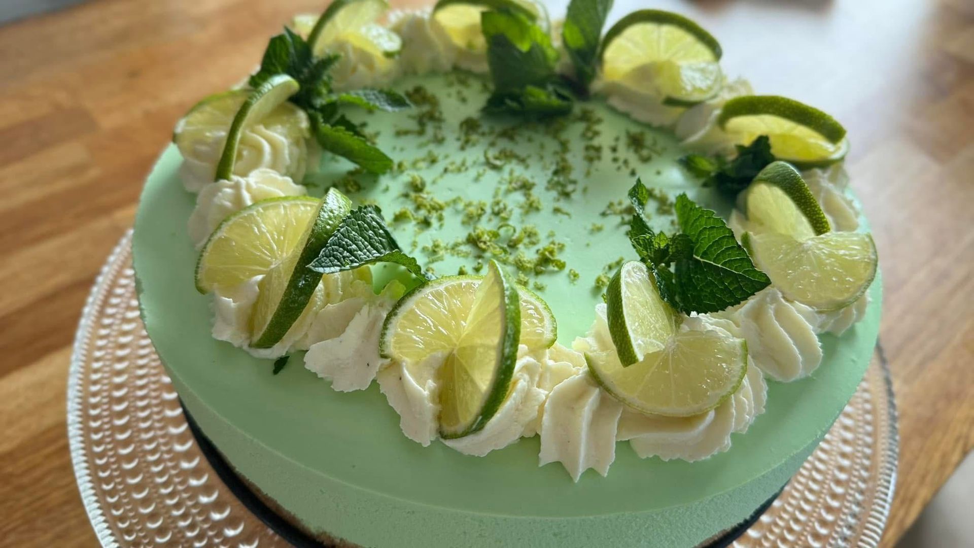 Limejuustokakku valkoisella kuorrutteella, limetiviipaleilla ja mintunlehdillä lasilautasella.