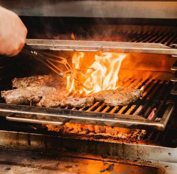 Pihvit grillataan metalliritilällä avotulen päällä; käsi käyttää pihtejä.
