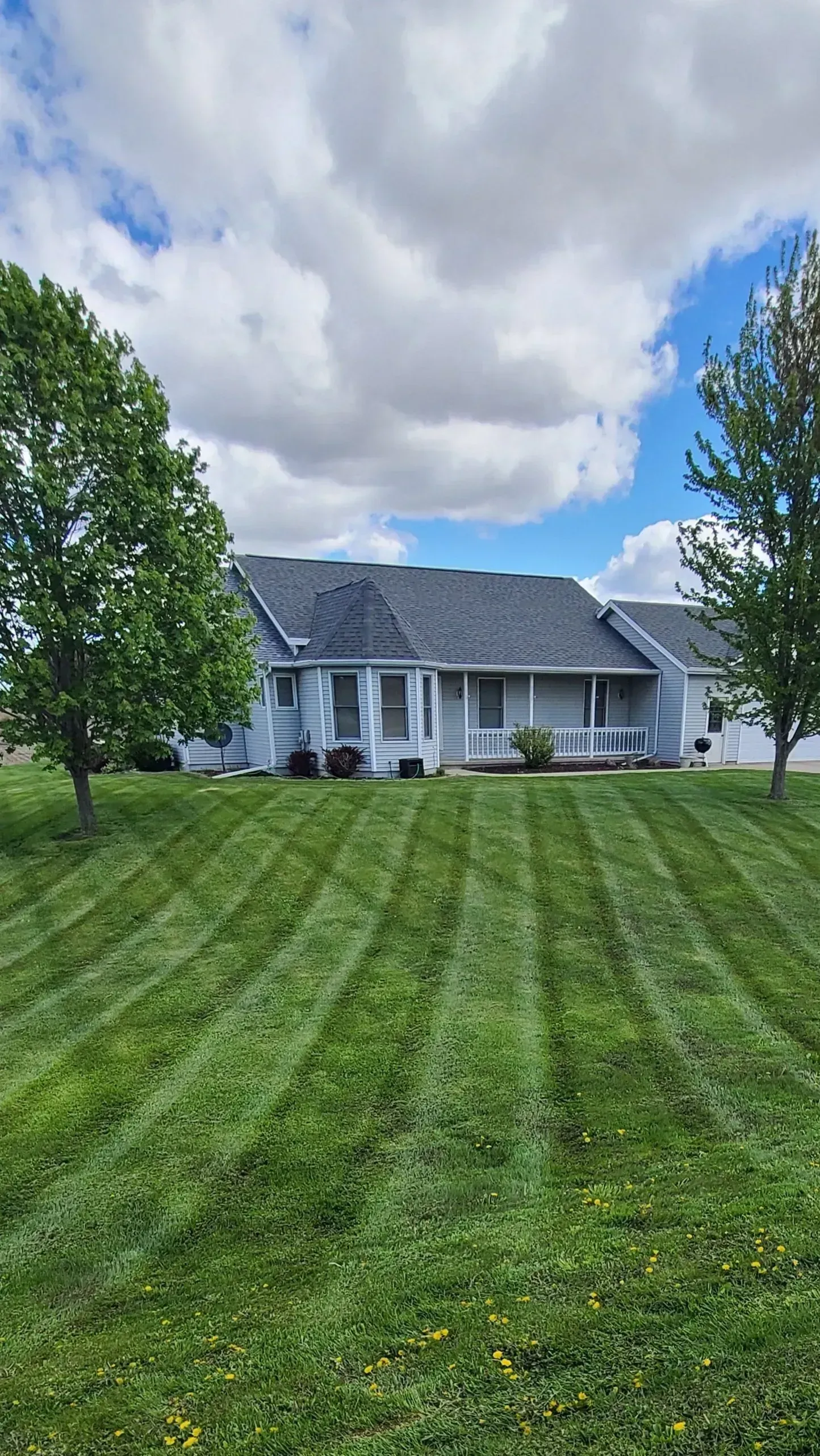 Suburban house with white siding and gray roof, seen across a striped green lawn under cloudy sky