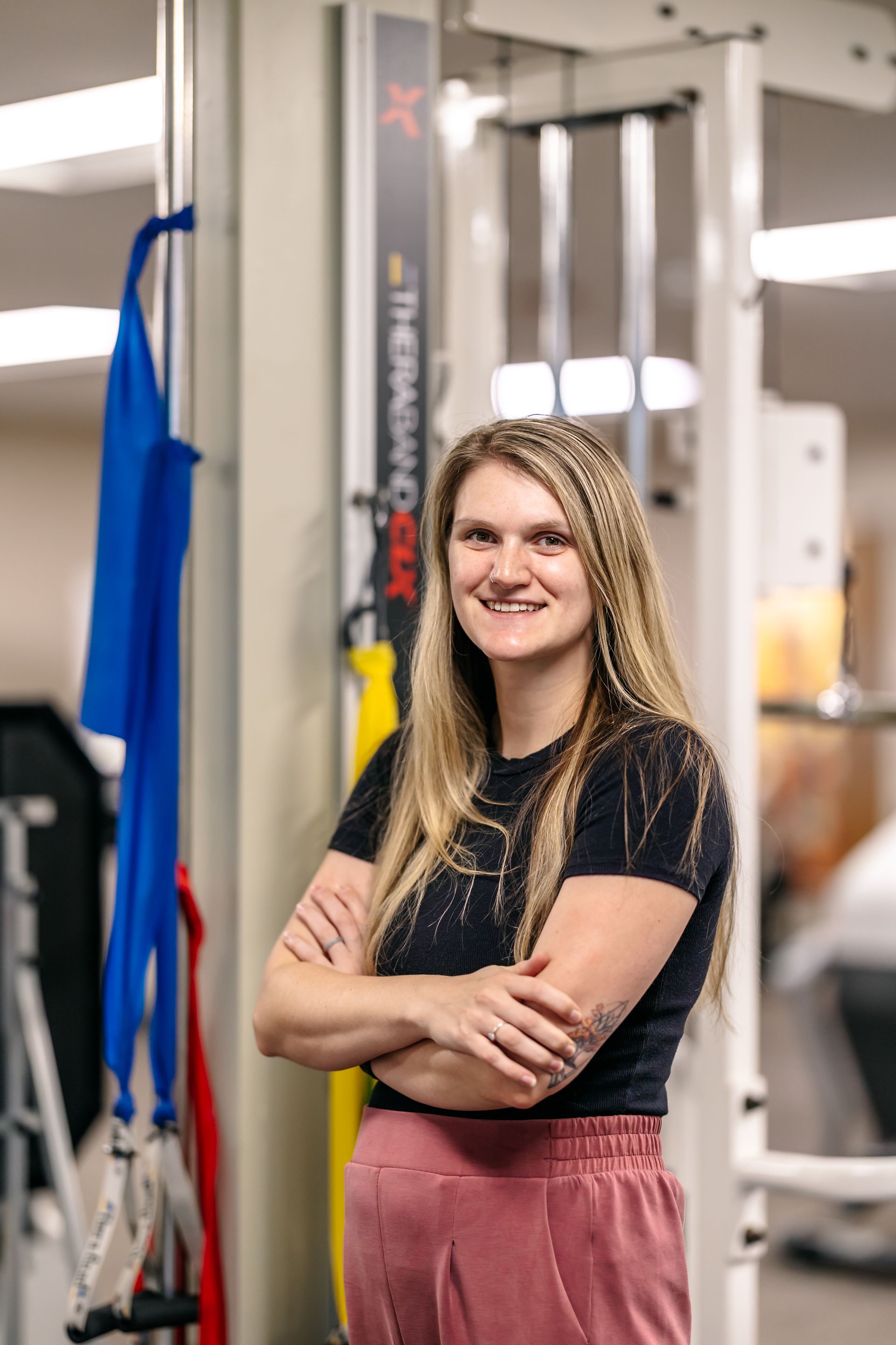 Woman standing with arms crossed in a gym, smiling beside exercise equipment.