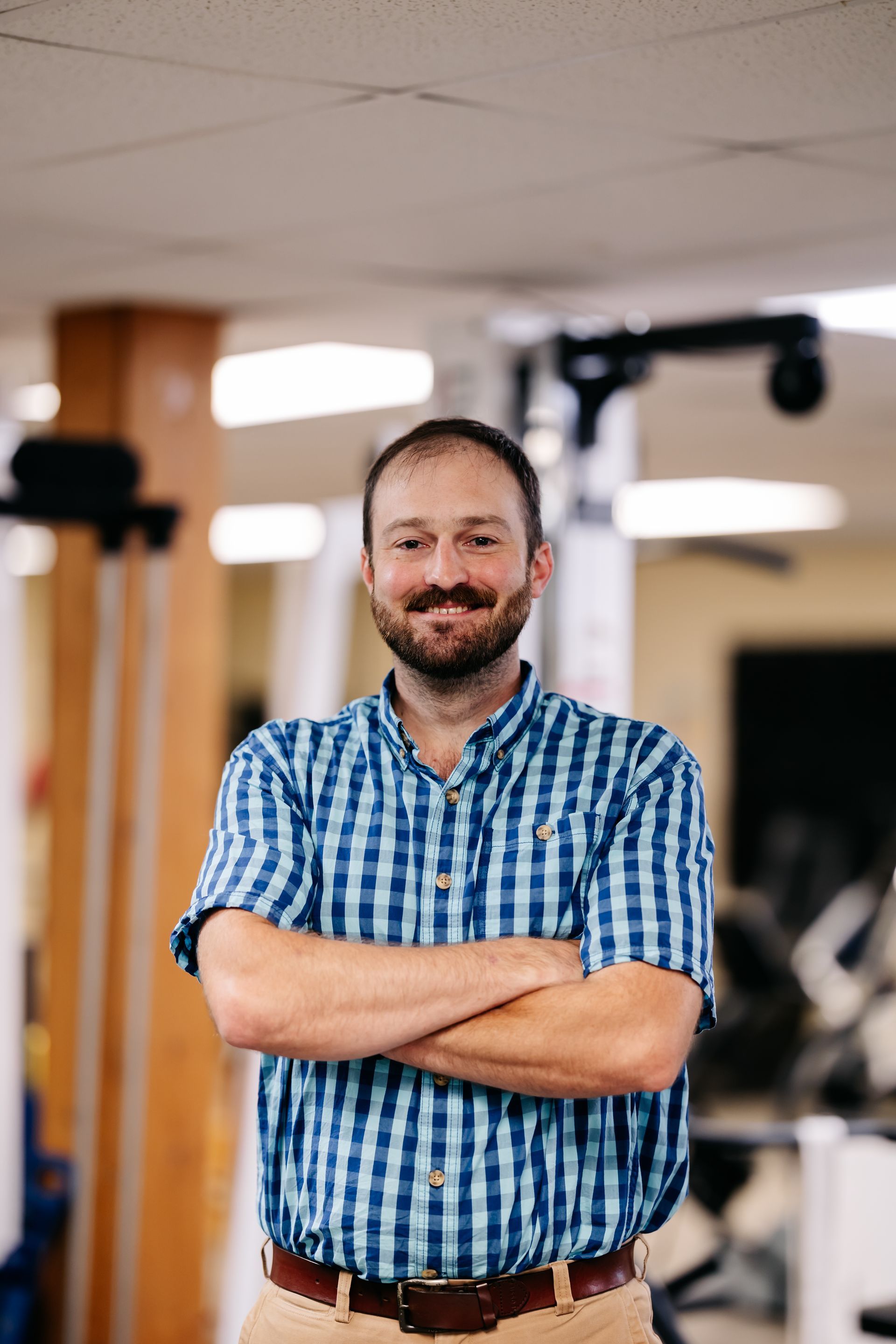 Smiling man with arms crossed in a blue plaid shirt standing in a gym.