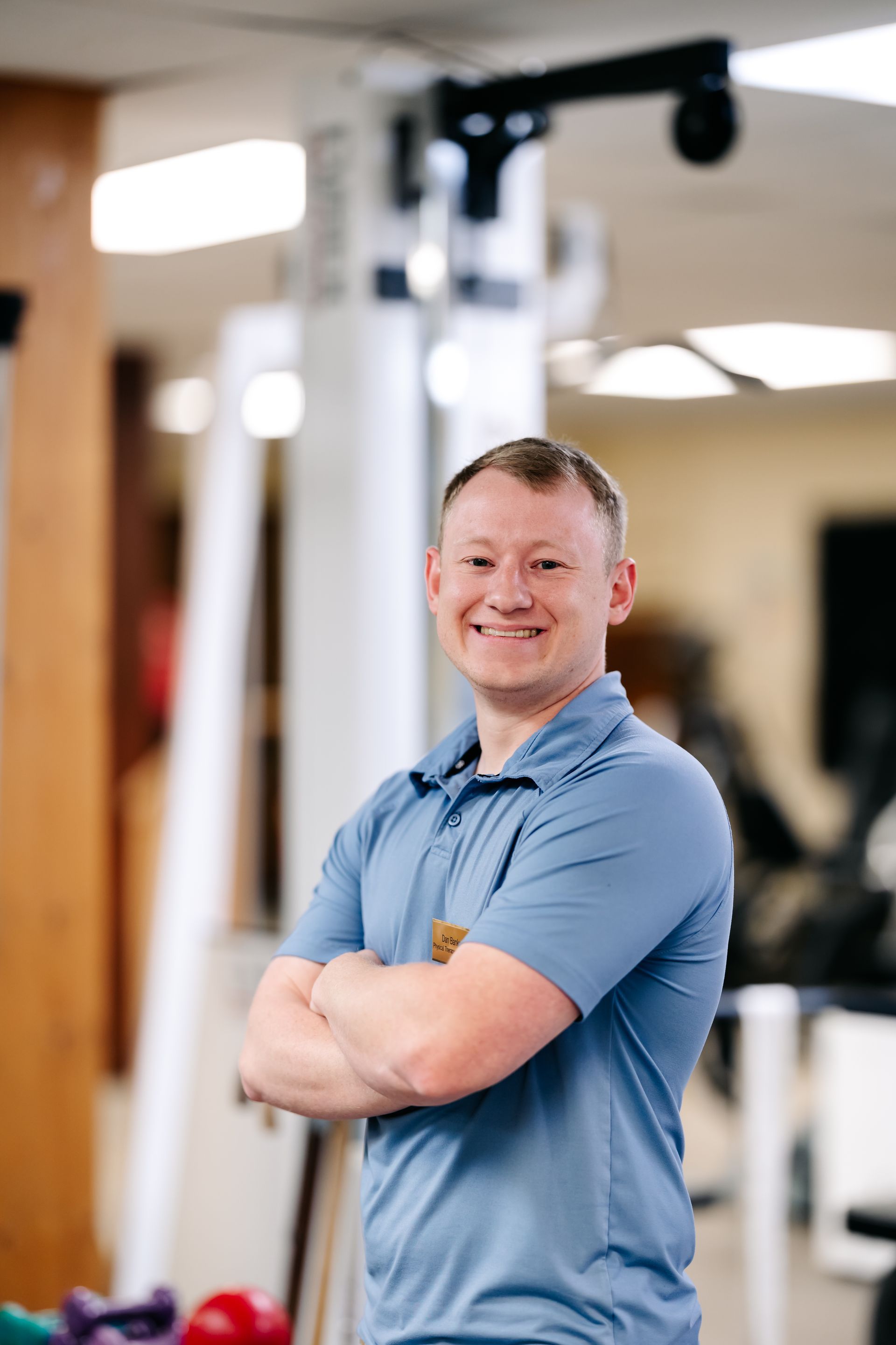 Smiling man in a blue polo stands with arms crossed in a gym.