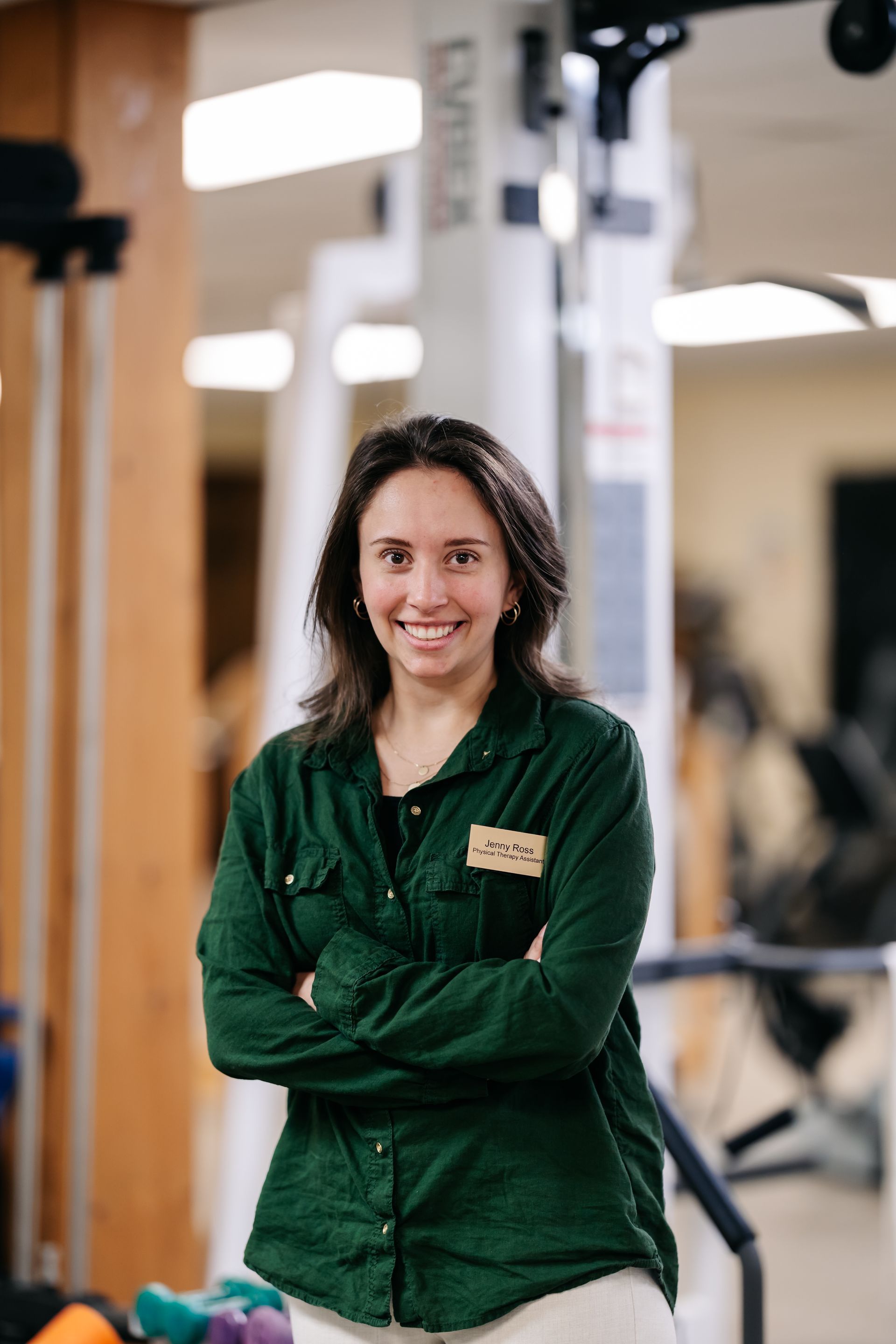 Woman in green shirt standing with arms crossed in a gym, smiling at the camera.