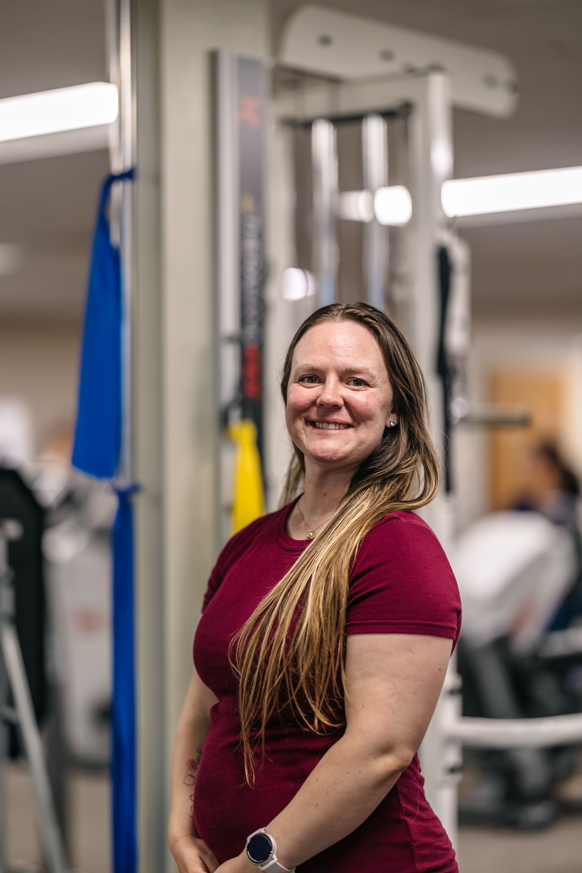 Woman smiling in a gym, standing beside exercise equipment in a maroon shirt.