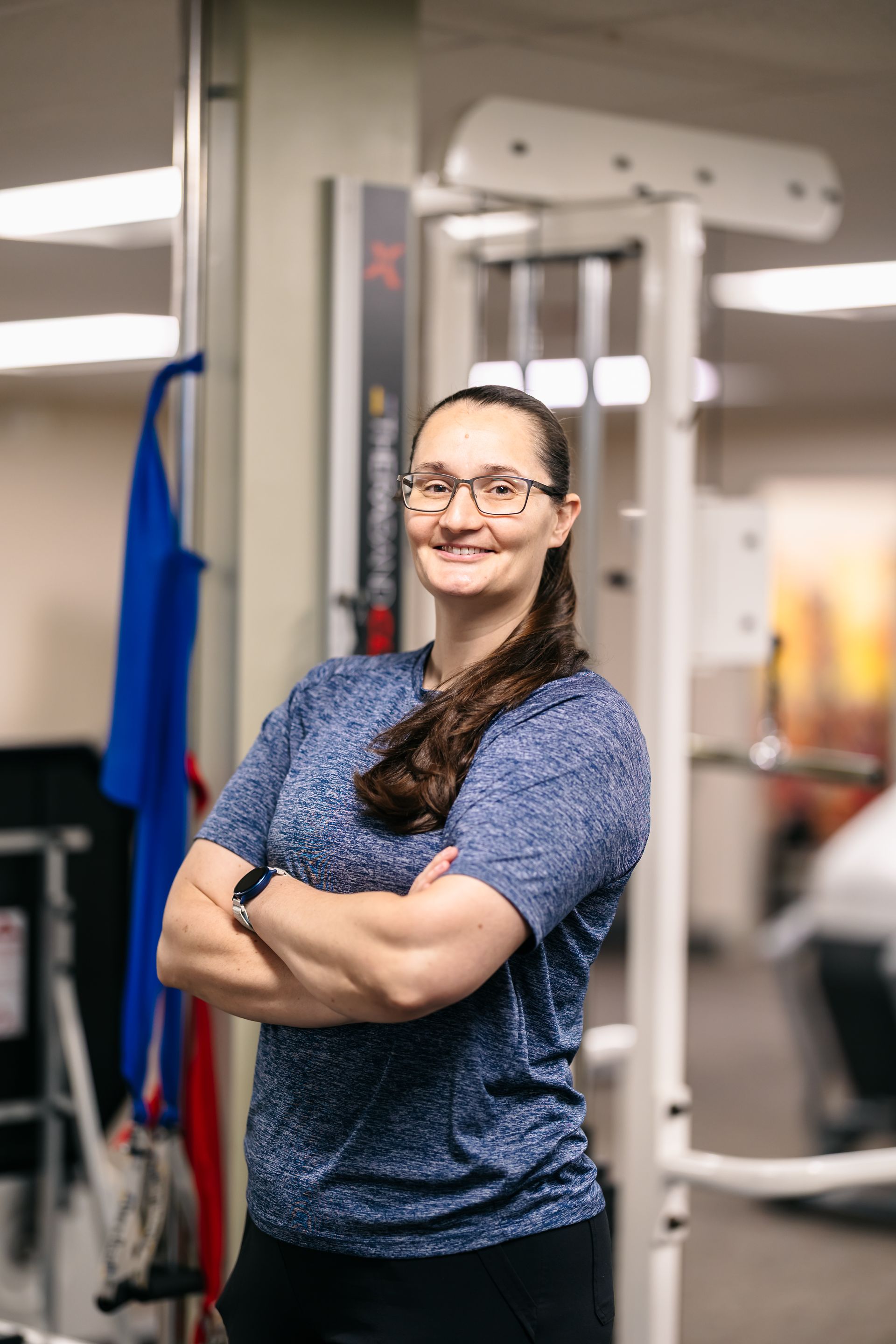 Woman standing with arms crossed in a gym, smiling beside exercise equipment.