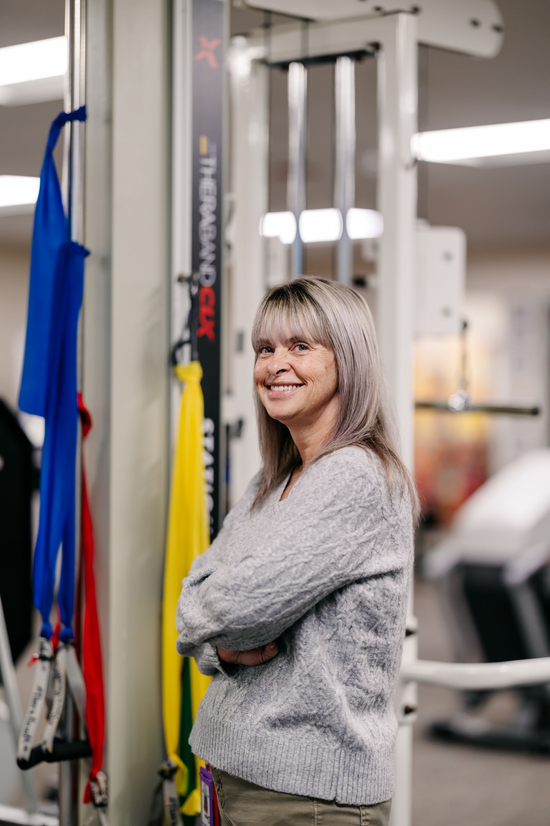 Woman smiling in a gym, standing beside exercise equipment with arms crossed.