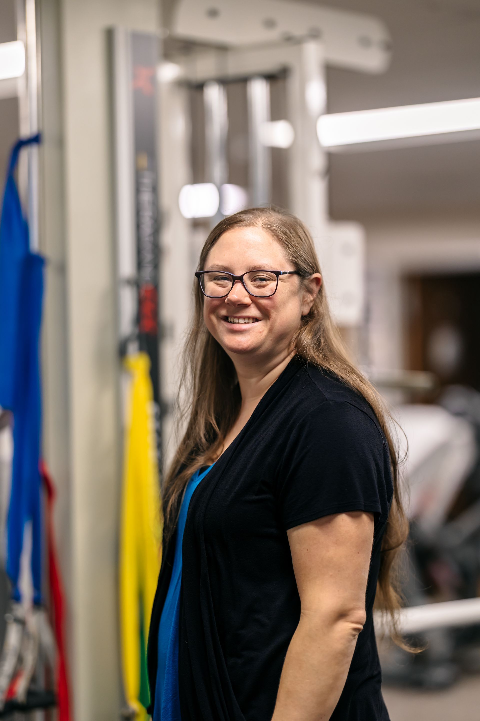 Smiling person with glasses in a gym, standing beside a cable machine and resistance bands