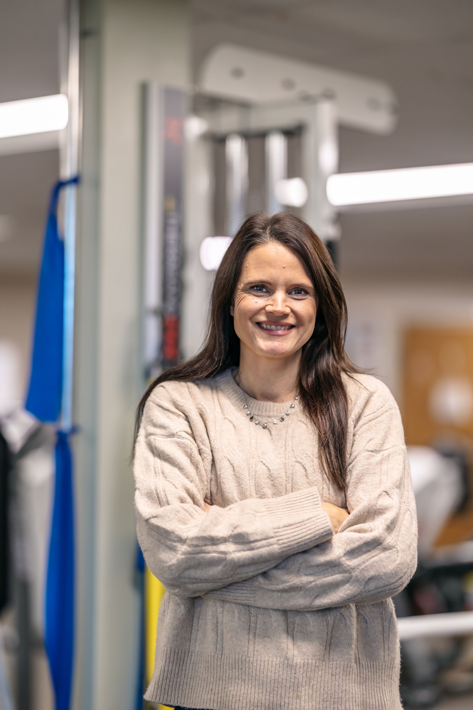 Smiling woman in a beige sweater standing with arms crossed in a gym or fitness room