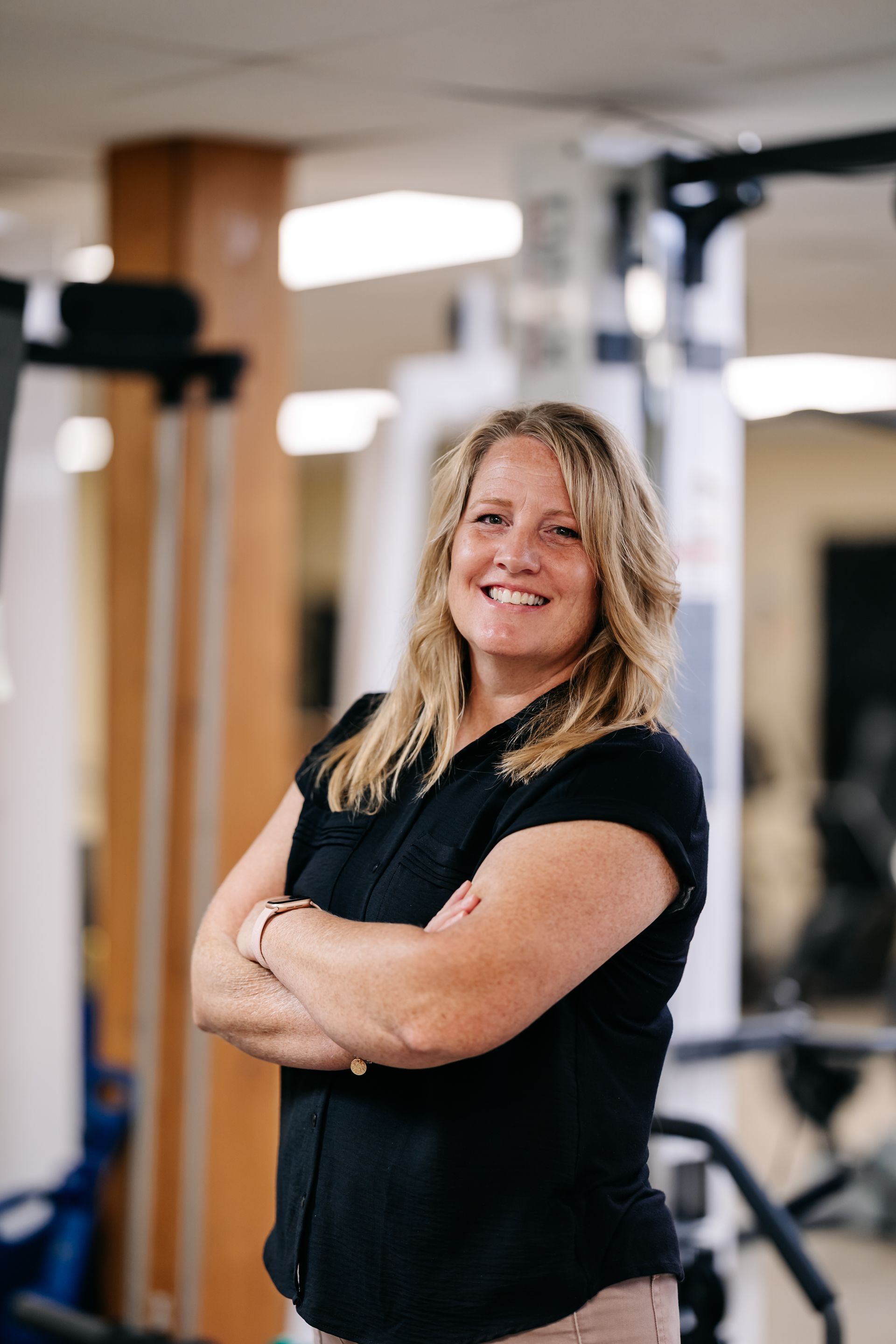 Woman standing with arms crossed in a gym, smiling in front of exercise equipment