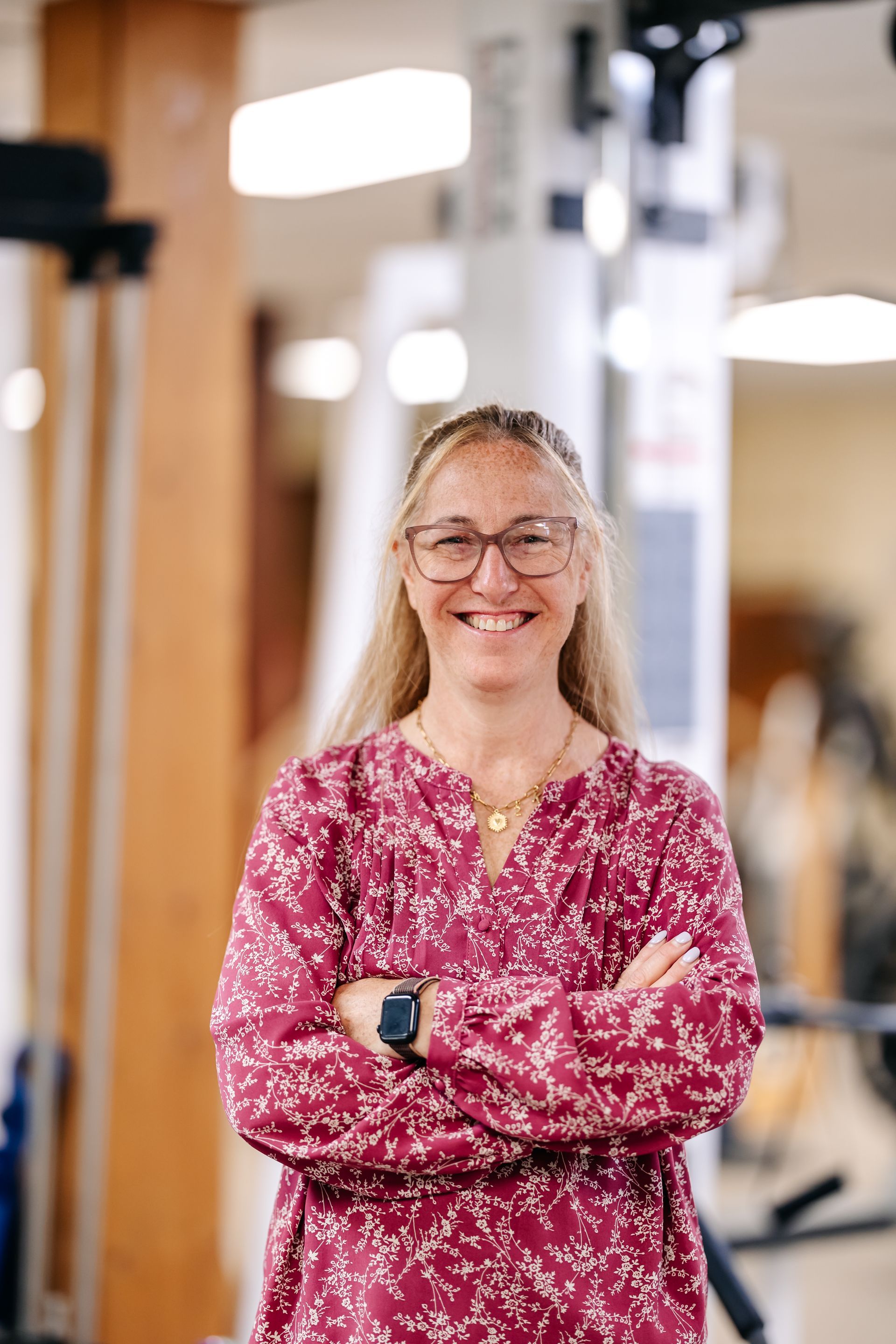 Woman in pink patterned top with arms crossed, smiling in a gym or fitness studio.
