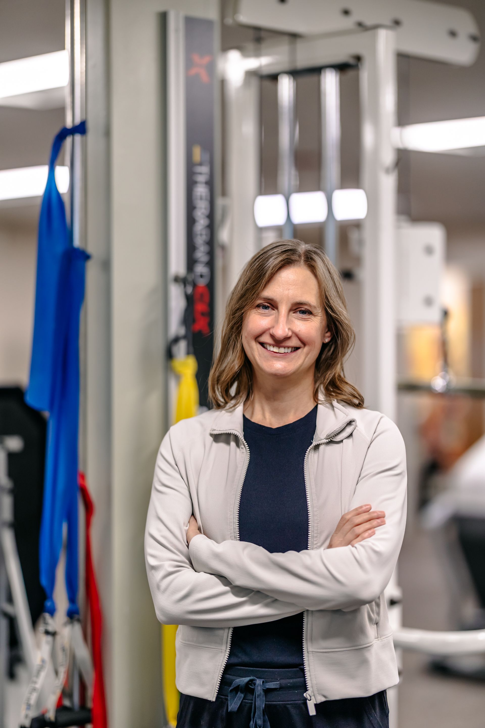 Smiling person with arms crossed in a gym, standing beside exercise equipment.