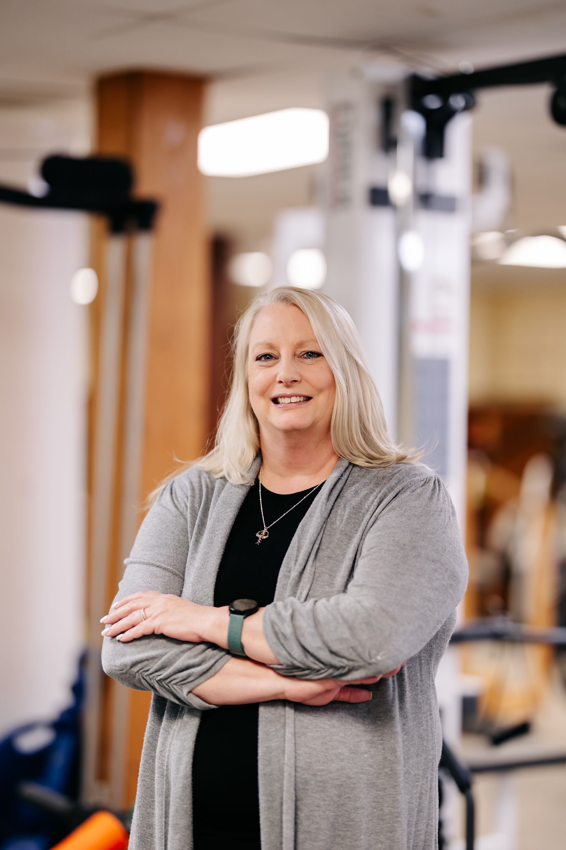 Smiling person standing with arms crossed in a gym, with exercise equipment in the background