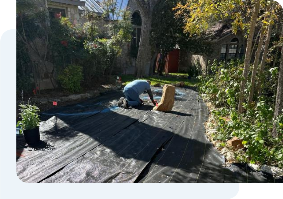 A man is kneeling on a black tarp in a garden doing a water sprinkler installation.