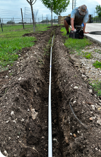 A man is working on a pipe in the dirt, repairing water sprinkler systems in the San Antonio area.