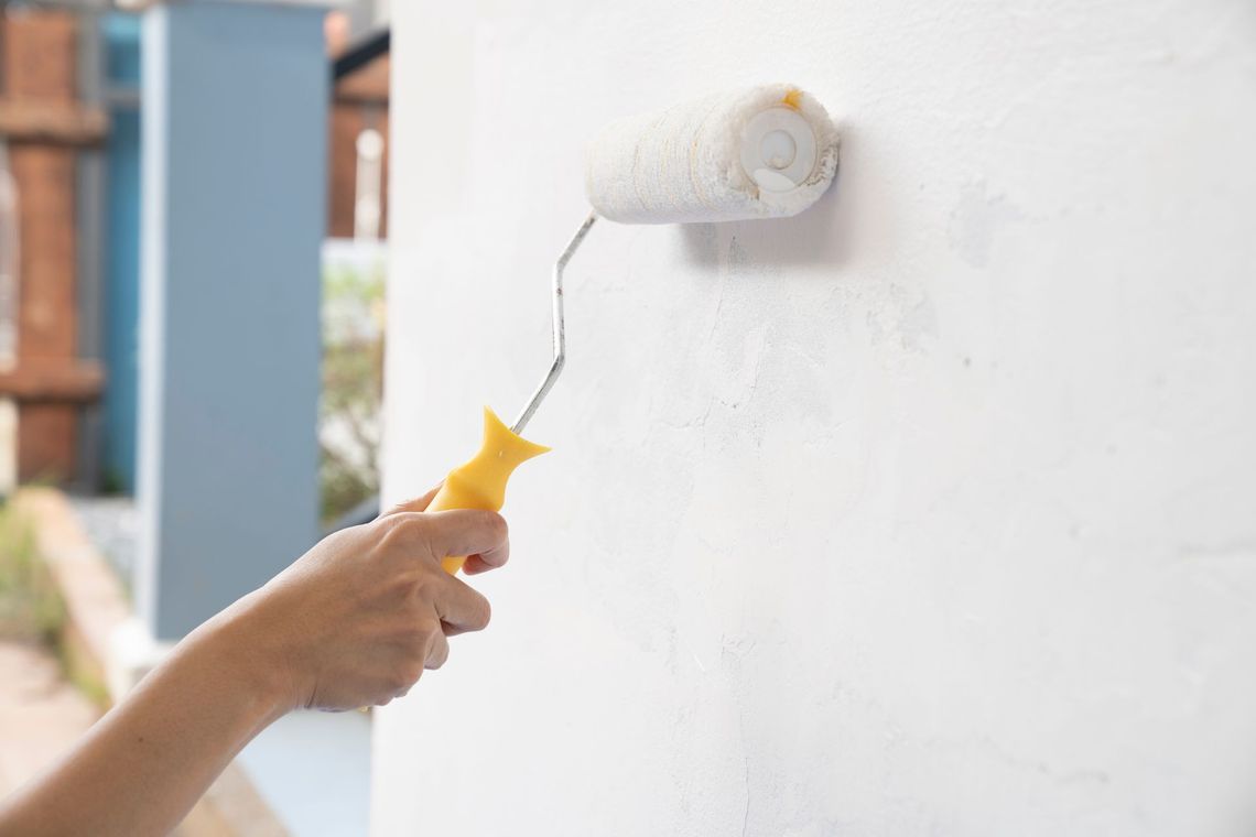Person painting a white wall with a paint roller; yellow handle, exterior setting.