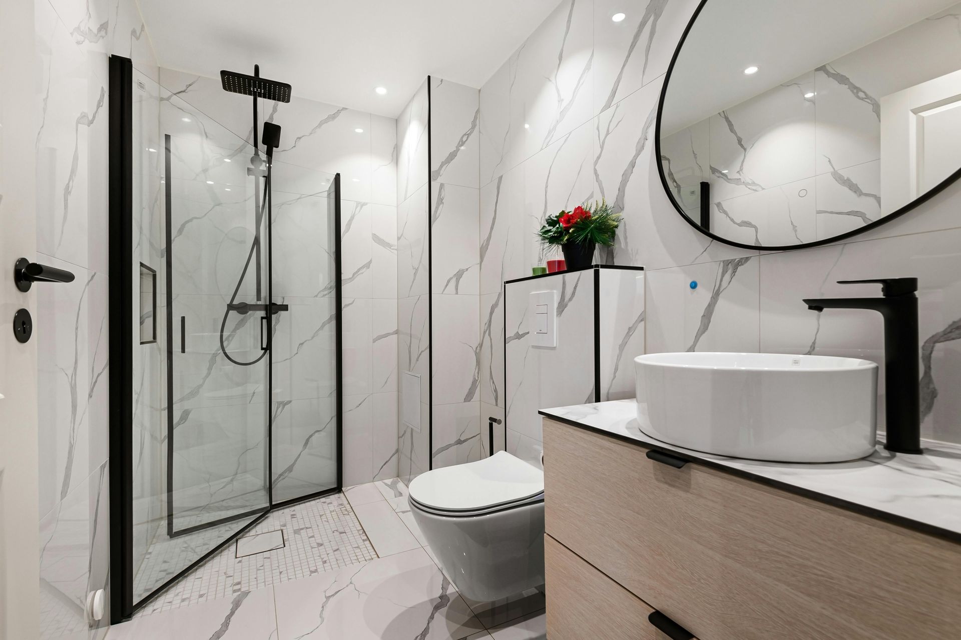 Modern bathroom with marble walls, glass shower, toilet, vanity, and round mirror