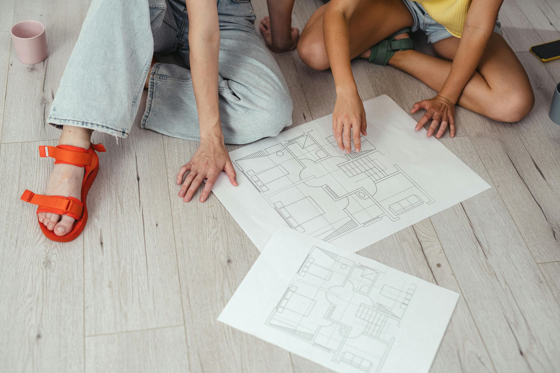People sitting on a light floor reviewing floor plan drawings, one wearing orange sandals