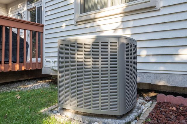 A grey outdoor air conditioning unit sits on a concrete pad next to the white siding of a house.