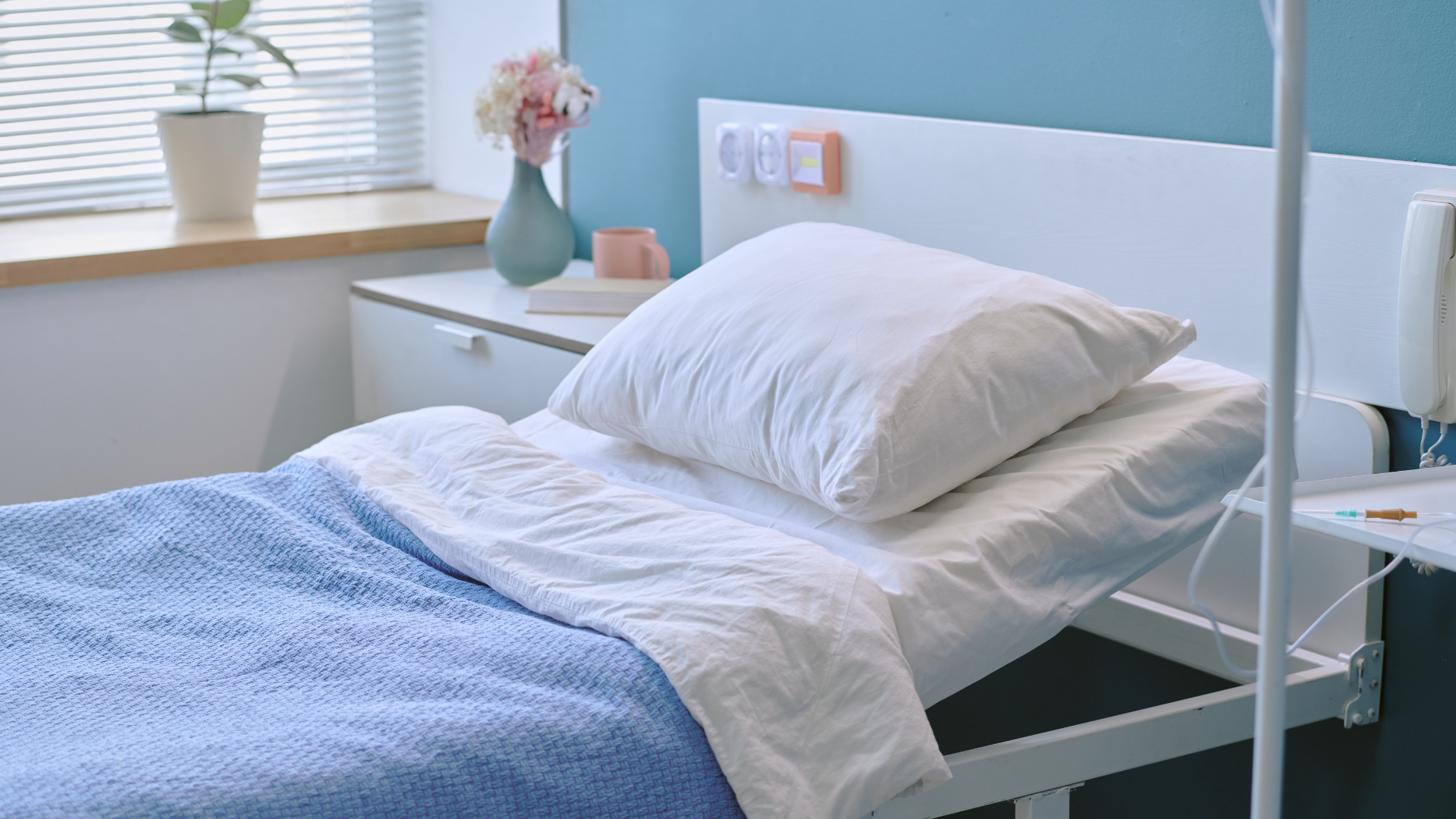 A neatly made hospital bed with a blue blanket, white pillow, and a small flower vase on a bedside table in a blue room.