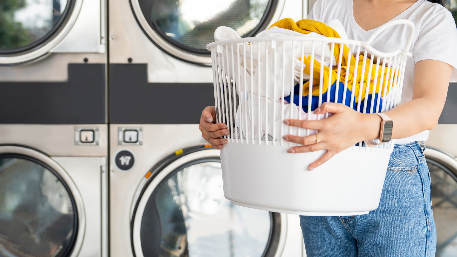 A person holding a white basket full of laundry in front of commercial washing machines at a laundromat.