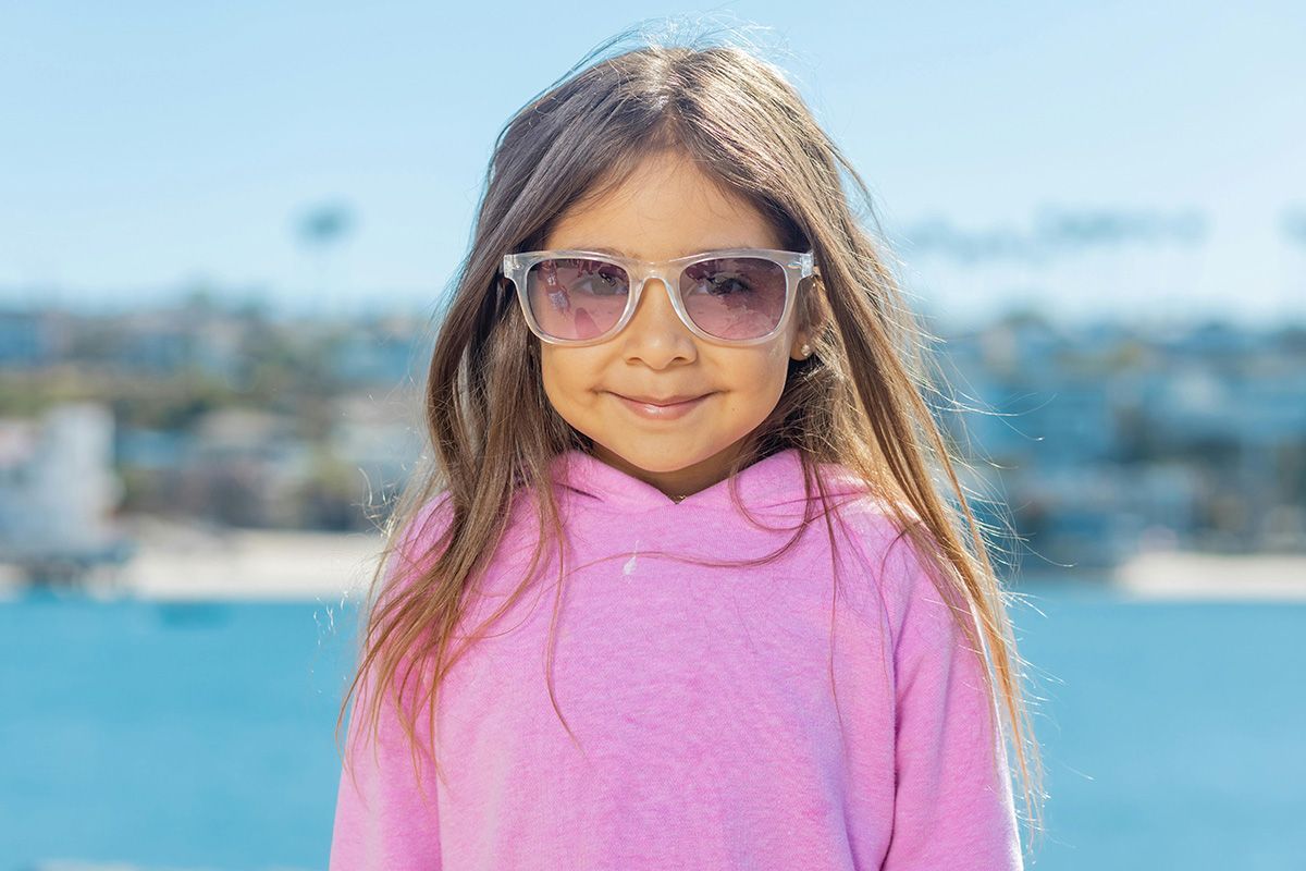Child in a pink sweater and sunglasses smiling outdoors with a blurred waterfront background