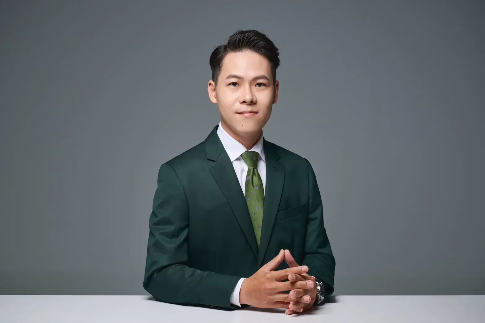 一名身穿深綠色西裝的男子坐在灰色背景的桌旁，雙手交握，看著鏡頭。