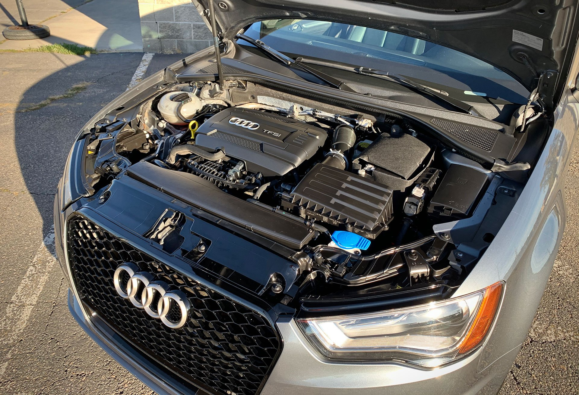An Audi car engine bay, top view. Black engine components, blue coolant cap, and silver car.