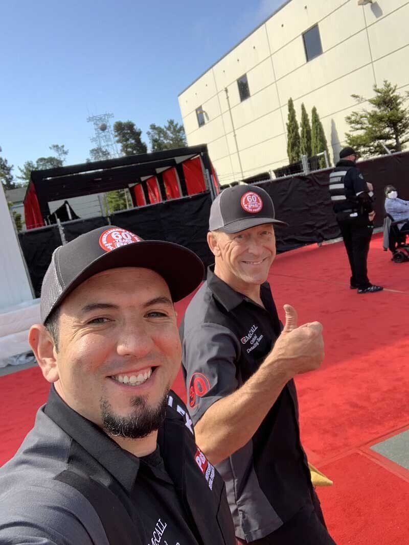 Two men in work attire smile on a red carpet. One gives a thumbs up. Sunny setting.