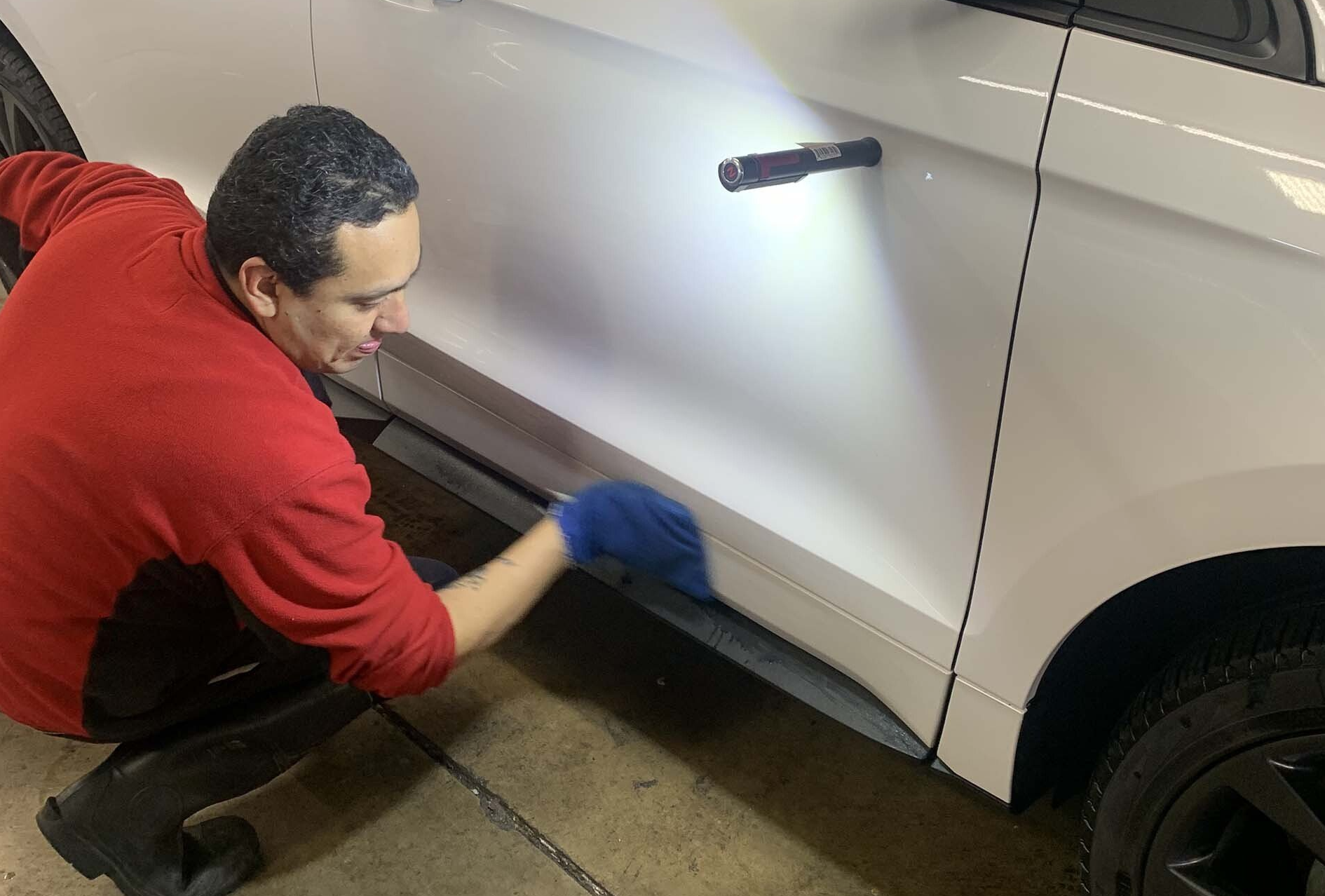 Man in red shirt cleaning a white car door with a blue glove, using a flashlight.